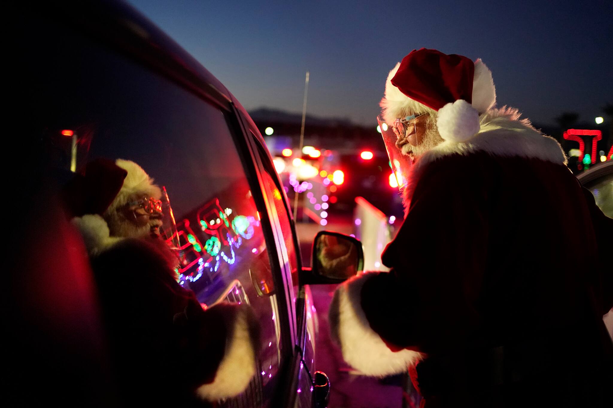 Charlie Bush, dressed as Santa Claus, wears a face shield as a precaution against coronavirus as he greets people waiting in their cars at Glittering Lights, a drive-thru holiday lights display, Thursday, Dec. 10, 2020, in Las Vegas. In this socially distant holiday season, Santa Claus is still coming to towns (and shopping malls) across America but with a few 2020 rules in effect. (AP Photo/John Locher)