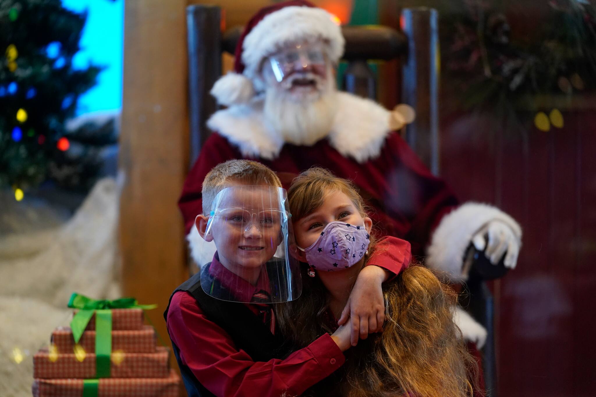 William Peargin, left, and Payton Peargin, right, both 8, pose for a socially distant photo with Santa Claus, who sits behind a sheet of plexiglass, at Bass Pro Shop in Rancho Cucamonga, Calif., on Dec. 4, 2020. In this socially distant holiday season, Santa Claus is still coming to towns (and shopping malls) across America but with a few 2020 rules in effect. (AP Photo/Ashley Landis)