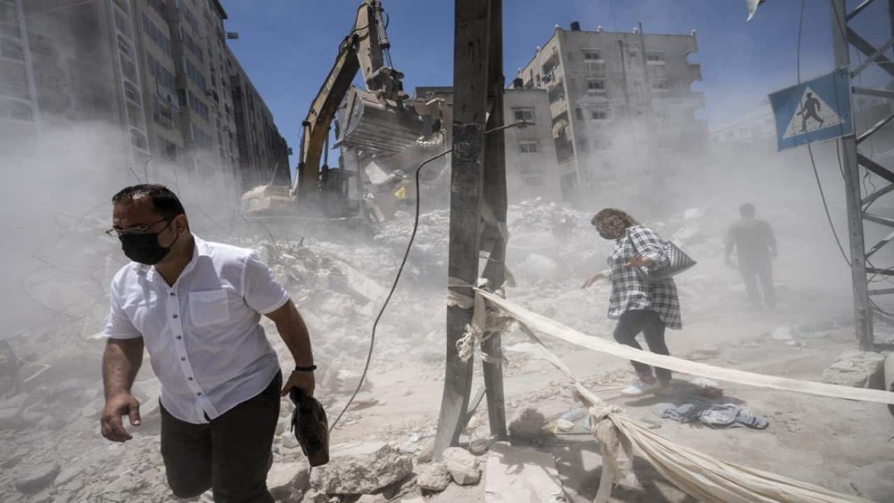 Pedestrians pass through clouds of dust as heavy construction equipment is used to sift through rubble to uncover valuables before it is transported away from the scene of a building destroyed in an airstrike prior to a cease-fire that halted an 11-day war between Gaza's Hamas rulers and Israel, Thursday, May 27, 2021, in Gaza City. (Image: AP)