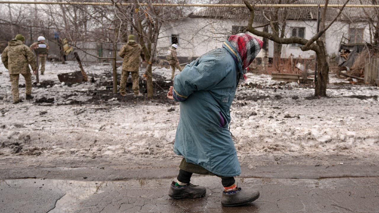 An elderly woman walks by as members of the Joint Centre for Control and Coordination on ceasefire of the demarcation line, or JCCC, survey a crater and damage to a house from artillery shell that landed in Vrubivka, one of the at least eight that hit the village today, according to local officials, in the Luhansk region, eastern Ukraine, February 17. (Image: AP)