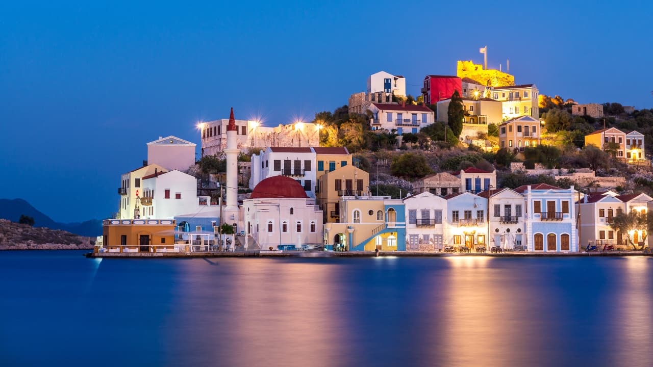 1. Kastellorizo – The Island That Whispers, Not Shouts Tucked at the southeastern edge of the Dodecanese archipelago, so close to the Turkish mainland it seems to lean toward it, Kastellorizo exists as a faded painting—soft-hued houses arranged around a horseshoe harbor, boats rocking like breath in sleep. This is not an island for spectacle but for quiet reawakening. One arrives here not to be entertained, but to remember stillness. (Image: Canva)