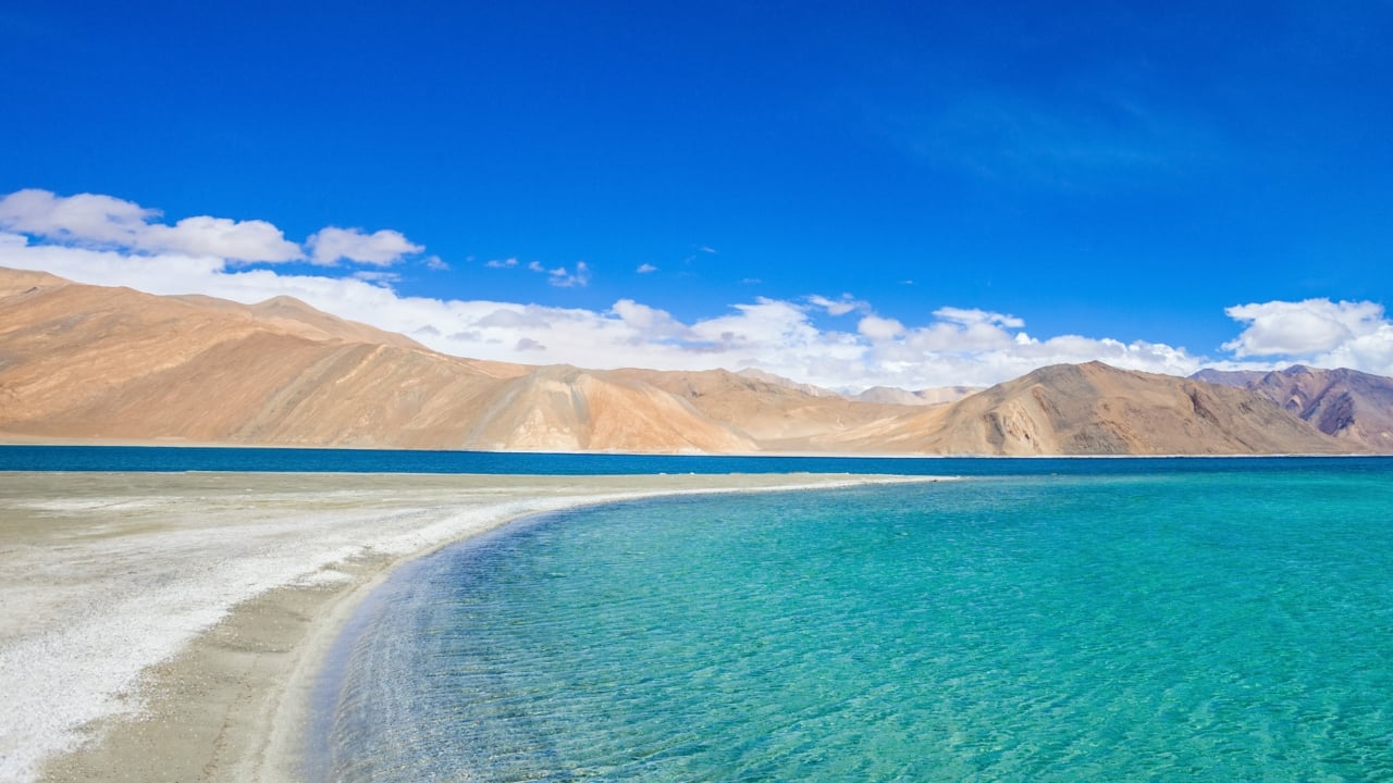 1. Pangong Tso, Ladakh Situated at an elevation of nearly 14,000 feet and straddling the Indo-Tibetan frontier, Pangong Tso is renowned for its constantly shifting colors. The lake’s palette moves effortlessly between slate blue, aquamarine, and olive green, set against a barren ridge that amplifies its visual drama. It is a minimalist masterpiece shaped by light and silence. (Image: Canva)