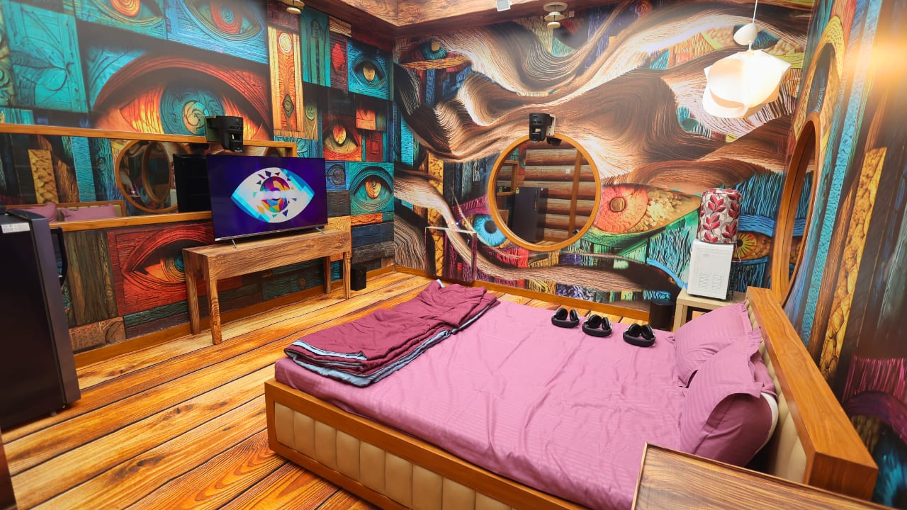 Private bedroom Bigg Boss 19 will feature a private bedroom area with wooden flooring contrasts with walls painted in vivid colours and covered in eye motifs, reminding housemates of constant surveillance. A large double bed dressed in purple adds vibrancy, while circular windows and rustic furniture maintain the natural theme. This space merges comfort with subtle psychological pressure. Private bedroom Bigg Boss 19 will feature a private bedroom area with wooden flooring contrasts with walls painted in vivid colours and covered in eye motifs, reminding housemates of constant surveillance. A large double bed dressed in purple adds vibrancy, while circular windows and rustic furniture maintain the natural theme. This space merges comfort with subtle psychological pressure.