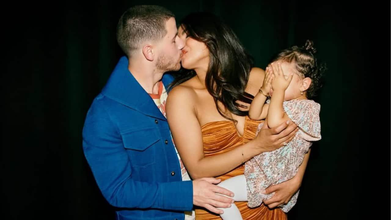 Laughter, love, and a hint of shyness  Priyanka Chopra and Nick Jonas kiss while holding their daughter Malti in this sweet moment. The couple is gorgeously dressed and exudes love, while little Malti covers her face in amusing embarrassment. This unscripted image perfectly combines humor, romance, and a tender family bond.