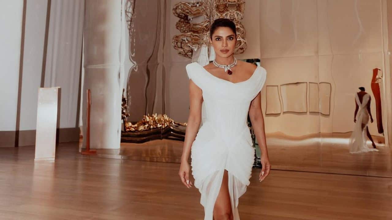 White gown and Bvlgari jewels Priyanka was photographed in a mirrored hall wearing a white off shoulder gown with a draped silhouette. The actress accessorized her dress with a deep red neckpiece.  The setting suggests an art gallery or event space, with abstract metallic art visible in the background reflections 