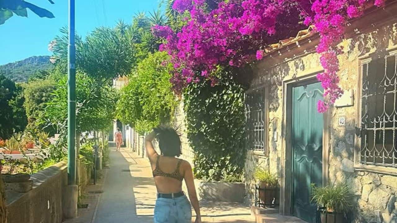 8. Tara Sutaria is photographed from the behind as she walks down a quaint, sunny alleyway. The scene is picturesque, with stone walls and a stunning canopy of bright pink flowers overhead.