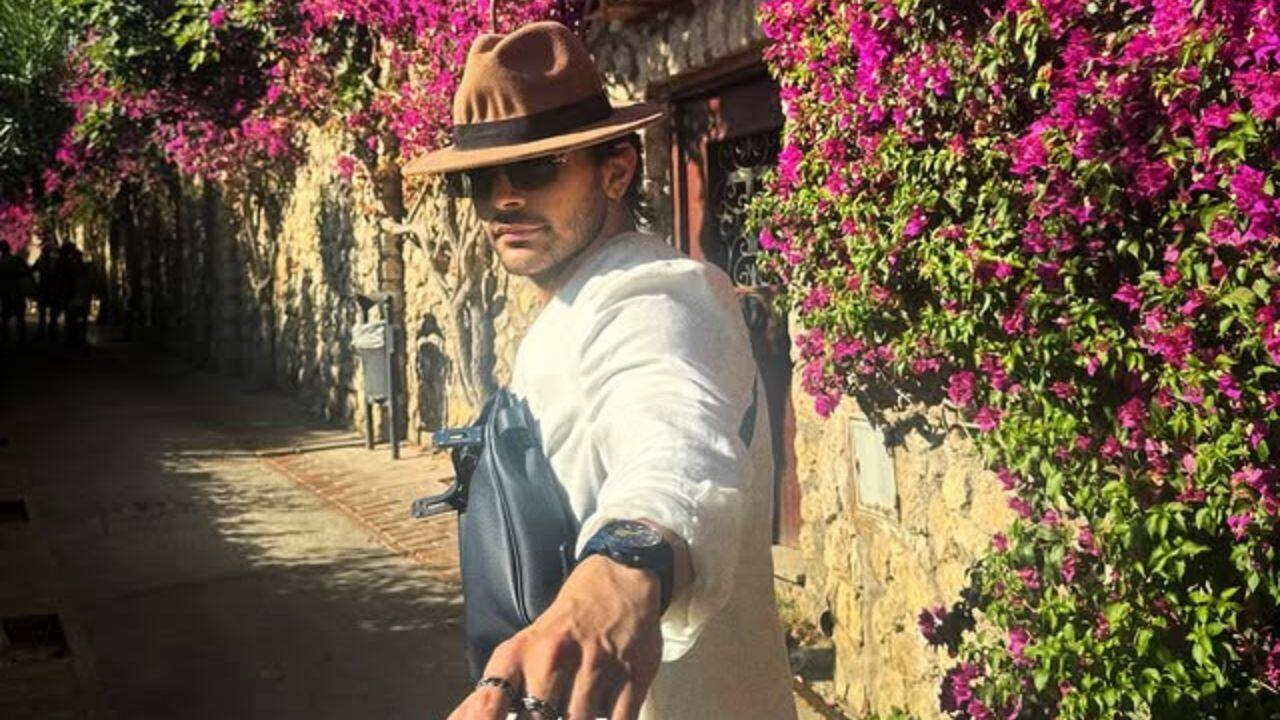 7. Veer Pahariya, dressed in a white suit and a fedora hat, is walking along a sunny stone pathway. The bright pink bougainvillea flowers in the background simple enhance the beauty of the picture. 