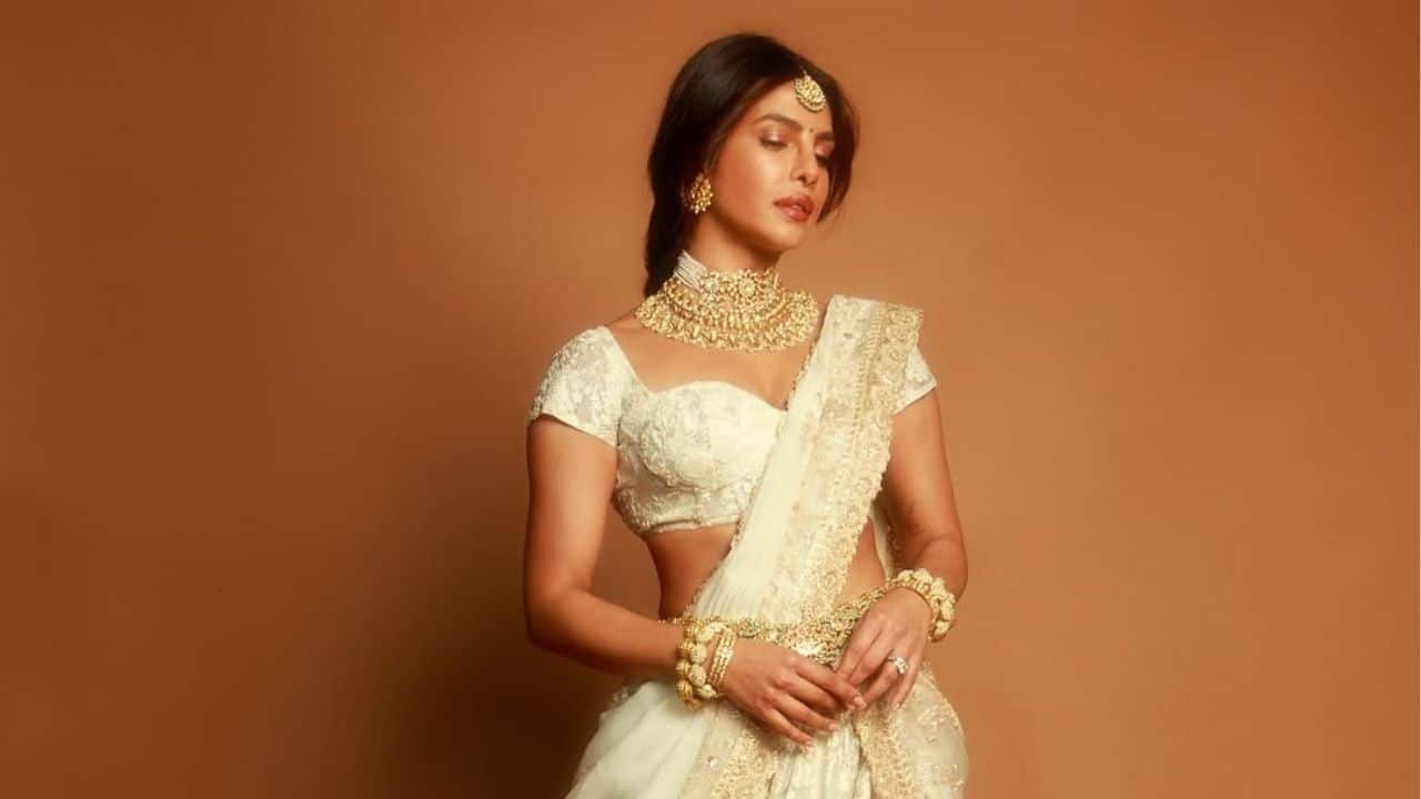 Layers of classic gold jewellery, such as a bold choker, matching earrings, and thick bangles, enhance Priyanka Chopra's appearance, while her soft makeup and well-com parted hair convey a subtle elegance. Priyanka appears ethereal, flawlessly expressing serenity and divinity with her eyes closed and her face serene.
