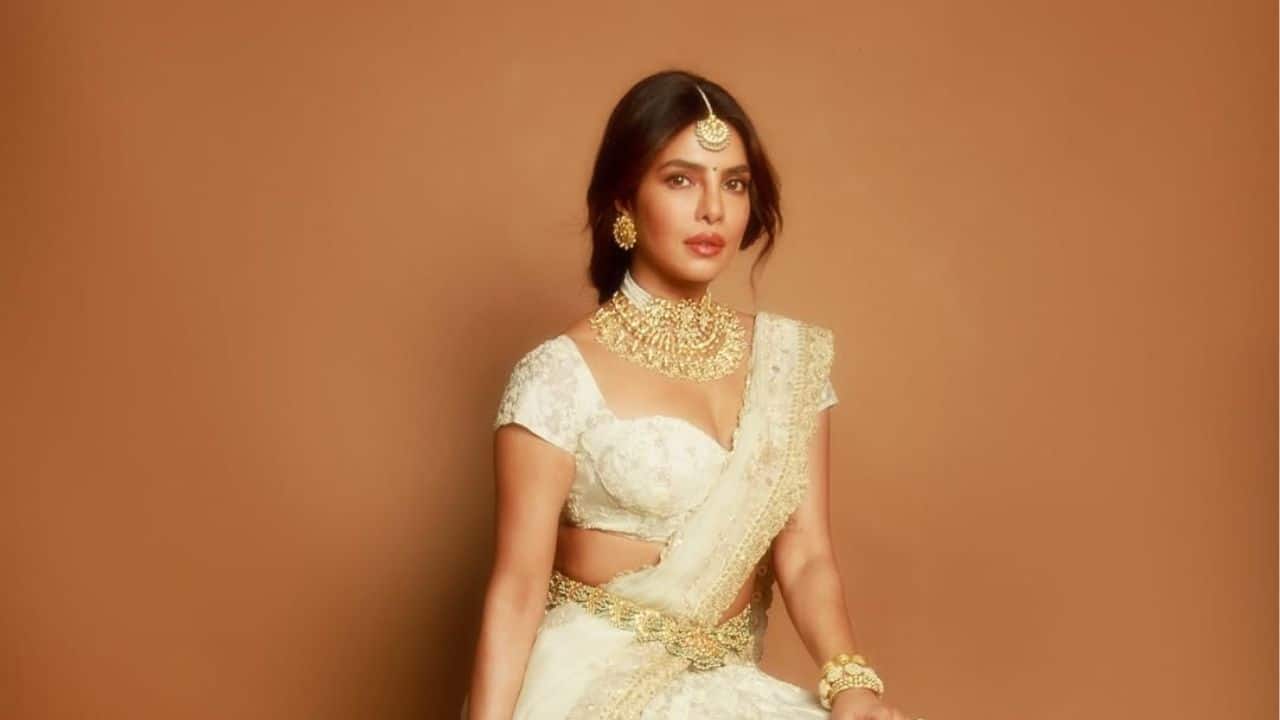 Sitting gracefully in an ivory saree with elaborate gold embroidery, Priyanka Chopra exudes divine grace and royal poise. Her delicate makeup, which is accentuated by warm tones and a delicate pink lip, accentuates her ethereal beauty, and her hair is styled in a clean centre part with soft waves framing her face.