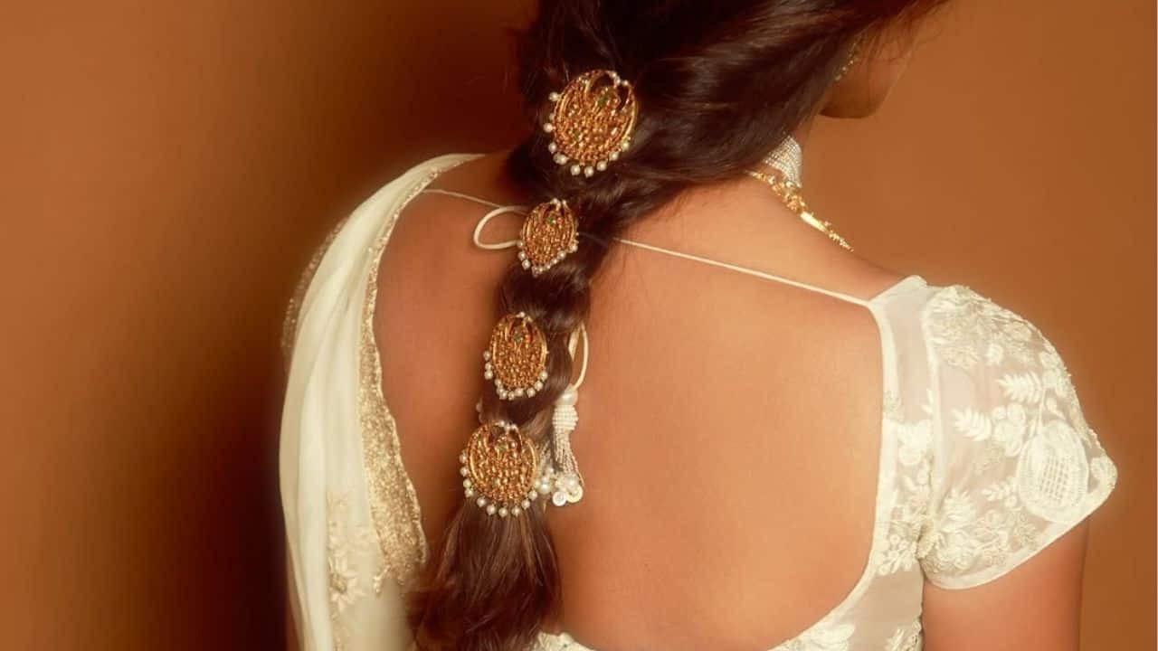With traditional golden jadanagam hair accessories delicately encrusted with pearls and green embellishments, Priyanka Chopra's intricately styled braid is beautifully captured in this close-up. Her embroidered ivory blouse and saree are exquisitely complemented by the intricately crafted ornaments, which combine classic elegance with sophisticated sophistication. Her heavenly, goddess-like appearance from the Varanasi event is completed by the delicate lighting, which accentuates the lustre of the jewellery and the richness of her hair.