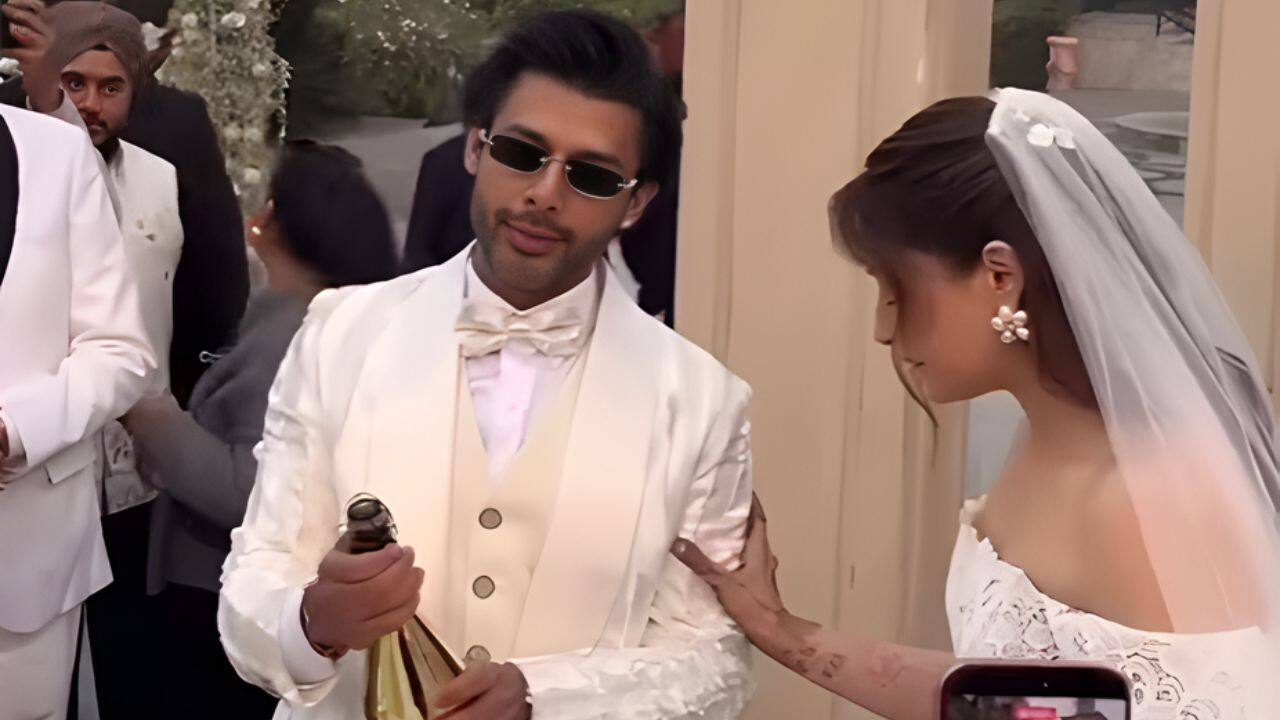 Stebin Ben and Nupur Sanon share a candid moment during their wedding festivities. Stebin looks sharp in a white tuxedo and dark sunglasses, holding a champagne bottle. Nupur, elegant in her white lace bridal gown and veil, stands beside him, resting a hand on his arm.