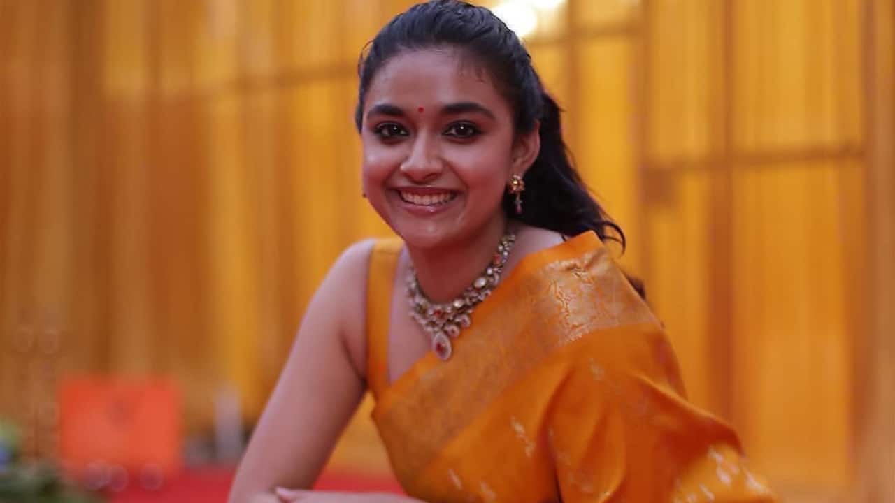 Vibrant Silk Glam Keerthy Suresh drapes a strappy halter-neck gold blouse elegantly over a vivid orange-gold Kanjeevaram silk saree with elaborate zari flower designs and a glittering pallu. Her accessories, which capture festive South Indian elegance, include a red bindi, matching bangles, stud earrings, a huge gold choker necklace, and her hair arranged in free waves with a side split.