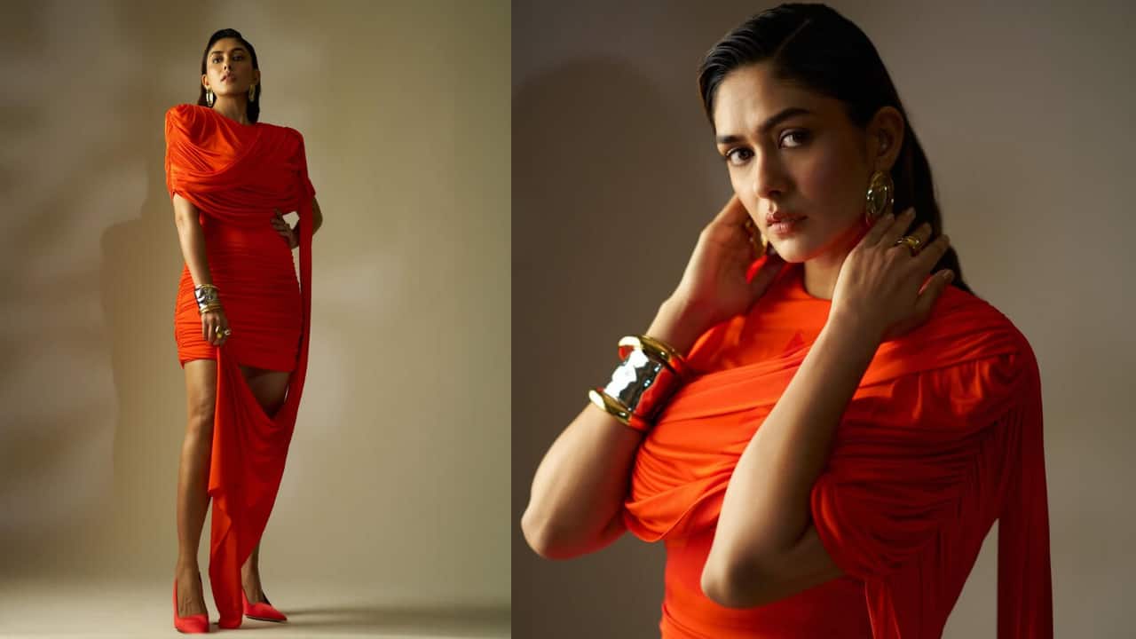 Perfect in crimson This vibrant orange-red mini dress is all about dramatic draping. The garment features intricate ruching and a bold, asymmetrical fabric trail that adds fluid movement. Styled with statement gold bangles and matching red pumps, the look is a powerful contemporary take on Grecian silhouettes, highlighting her sharp, editorial posing.