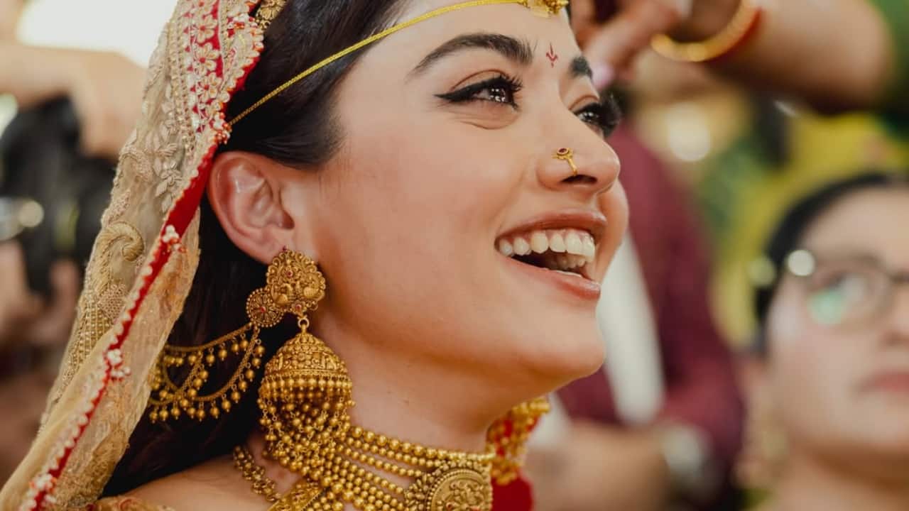 A close portrait of Rashmika captures her glowing smile, framed by heavy temple jewellery and a red bridal sari. The sacred markings on her forehead and delicate nose ring complete the traditional look. She appears radiant, confident and deeply present in the moment.