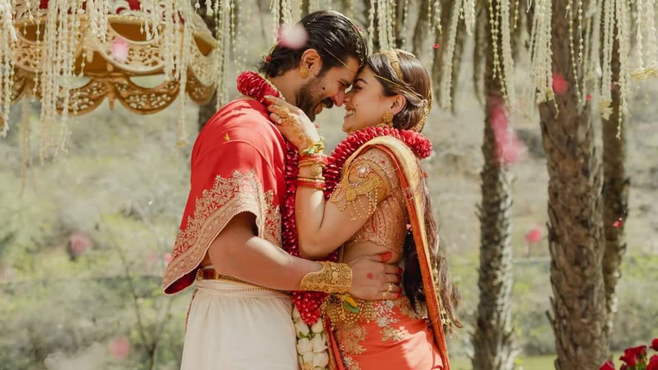Under cascading white flowers and drifting rose petals, the couple embraces during a dreamy outdoor moment. Surrounded by nature and soft smoke effects, their red and ivory ensembles glow in natural light. The image feels cinematic, almost like a still from a grand romantic epic.
