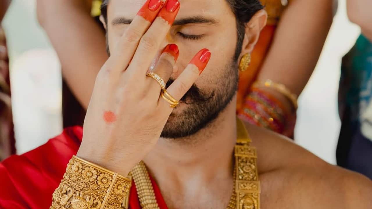 A striking close-up shows Vijay with alta-stained fingers partially covering his face. Layered gold temple jewellery, a red drape and sacred threads complete the powerful look. The image reflects a groom fully immersed in ritual, redefining masculinity through confidence and cultural pride.